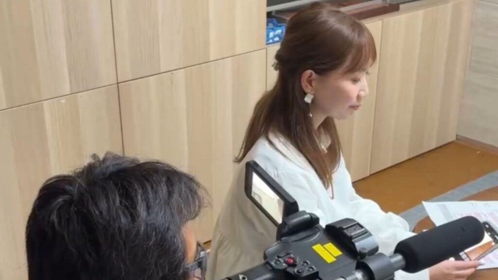 フリマ物販講師みき先生がテレビ番組の収録現場でマイクをつけ、スタッフと打ち合わせしながらフリマのテクニックについて説明している真剣な表情の様子